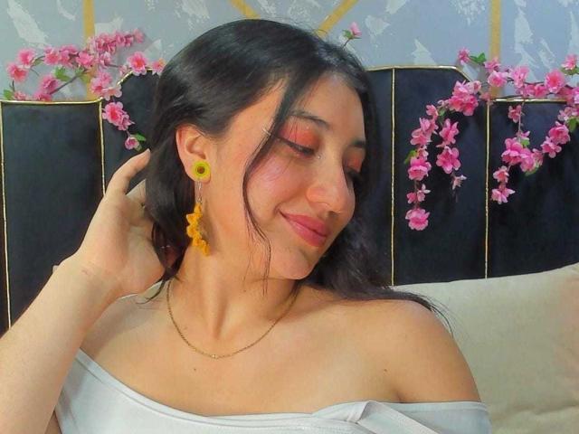 ArianaZahar with dark hair, yellow earrings, orange eyeshadow, off-shoulder white top, and subtle smile, hand in hair, pink flowers in background, looking down, soft lighting.
