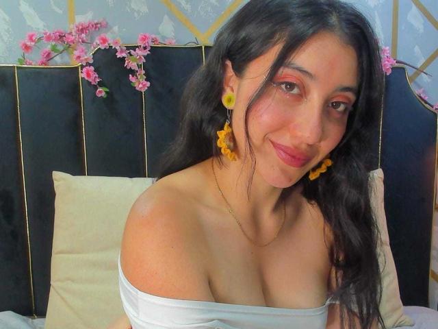 ArianaZahar with long black hair, yellow flower earrings, and a white off-shoulder top, showing her cleavage, smiling at the camera, pink eyeshadow, and yellow flowers in her hair.