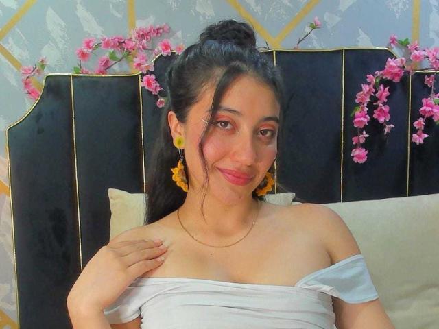 ArianaZahar with dark hair in a bun, yellow earrings, pink lipstick, off-shoulder white top, smiling, hand on chest, pink flowers behind black headboard.