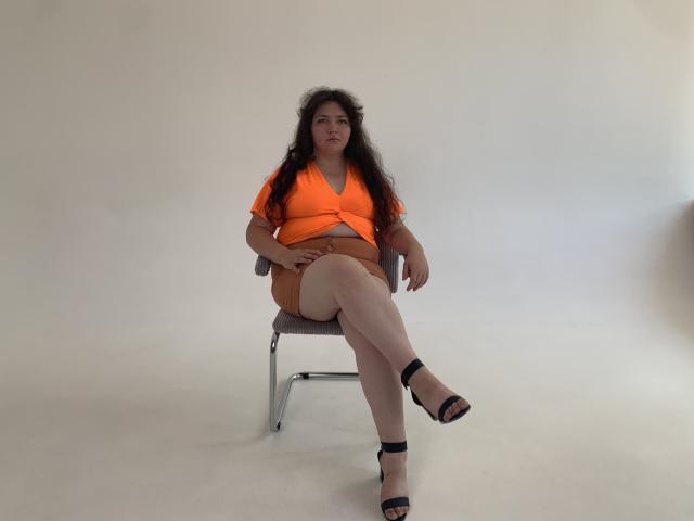 NeonilaLisova with curly dark hair, medium boobs, orange top, orange shorts, black strappy heels, sitting on a chair, legs crossed, white background, looking slightly down.