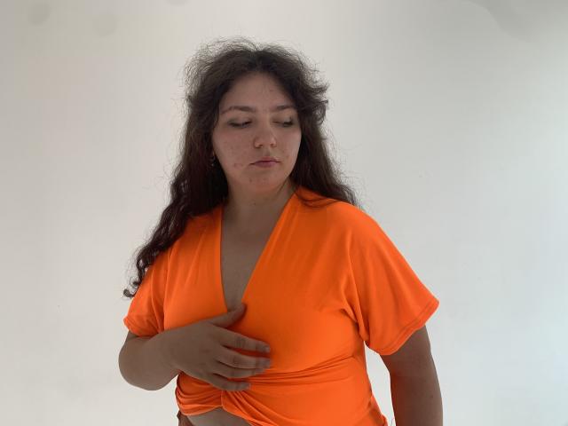 NeonilaLisova with messy dark hair, eyes closed, wearing an orange crop top, holding her tits with her right hand. Her face has a few small acne spots.