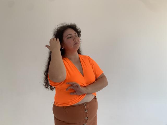 NeonilaLisova with curly black hair, orange v-neck top, and brown button-up pants, touching her head with one hand, eyes closed, slightly frowning, standing against a white wall.