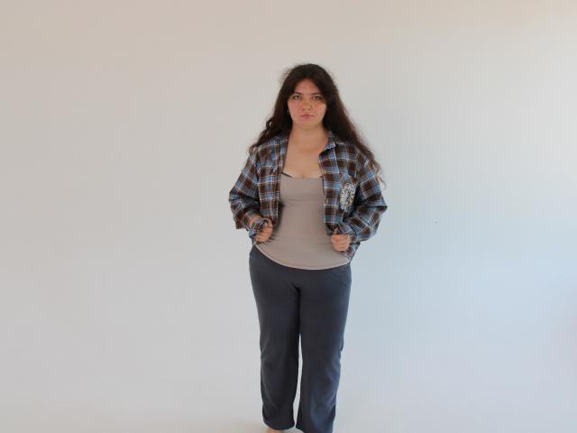 NeonilaLisova stands with hands in plaid shirt pockets, wearing a gray tank top and dark pants, long black hair, and a serious expression, showing small tits and a soft ass.