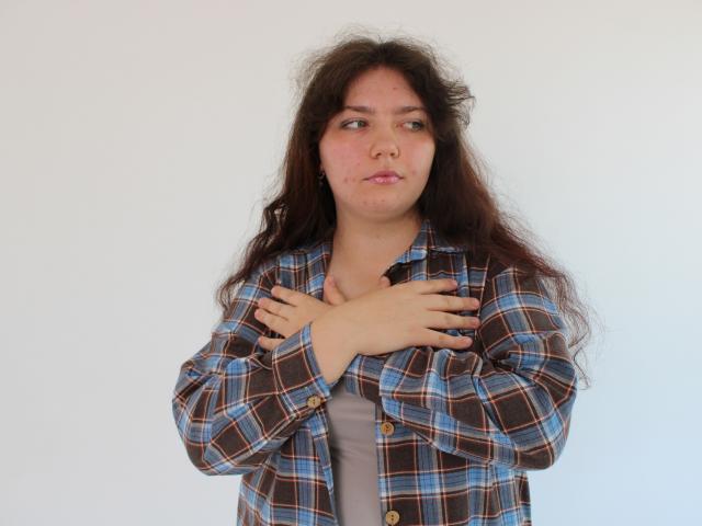 NeonilaLisova has long, messy brown hair, a slightly puffy face, and is wearing a blue and brown plaid shirt, crossing her hands over her chest.