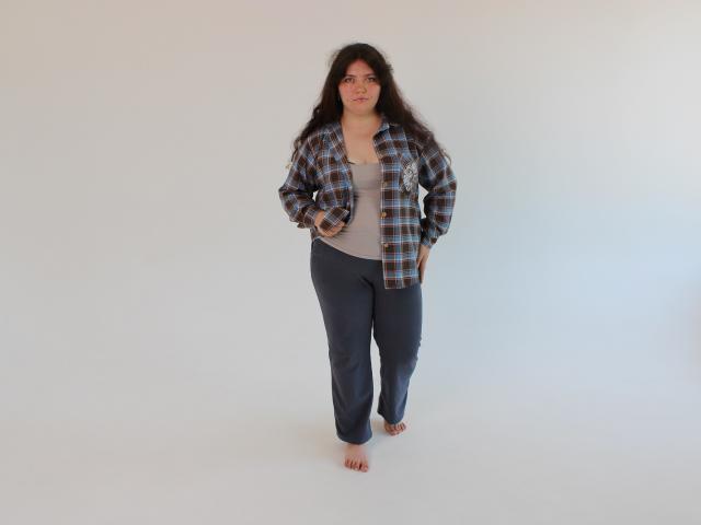 NeonilaLisova with long dark hair, wearing a plaid shirt, gray tank top, and dark pants, standing barefoot, hands in pockets, looking slightly to the side.