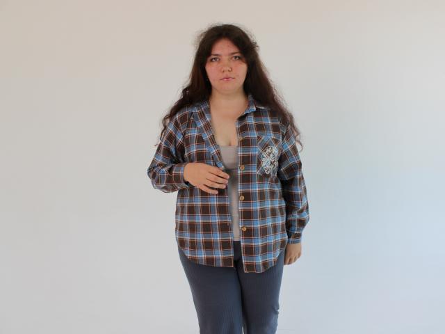 NeonilaLisova has long dark hair, wearing a blue and brown plaid shirt over a gray top, with dark pants. She's standing with one hand near her chest.
