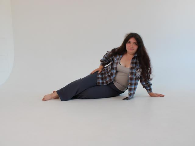 NeonilaLisova has long dark hair, wearing a plaid shirt, grey tank top, and blue pants, sitting on white floor, barefoot, with a relaxed, slightly seductive expression.