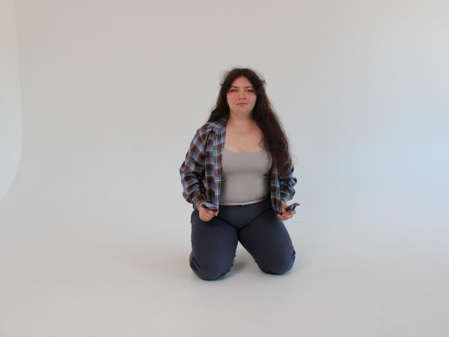 NeonilaLisova with long dark hair, wearing a plaid shirt, gray tank top, and dark pants, kneeling on white floor, holding keys, looking at camera with a neutral expression.