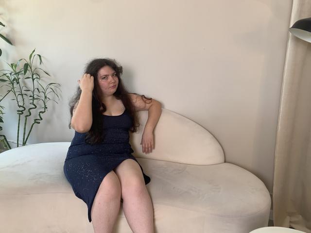 NeonilaLisova with long dark hair, wearing a dark blue dress, sitting on a white couch, hand in hair, looking at camera, slightly smiling, legs crossed, arms resting.