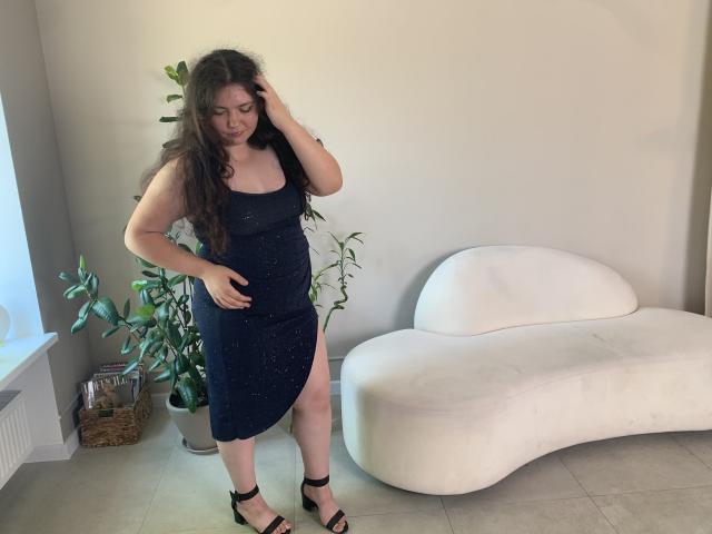 NeonilaLisova has long brown hair, wearing a dark blue dress with a thigh slit, black heels, and is standing with one hand in her hair, the other on her belly.