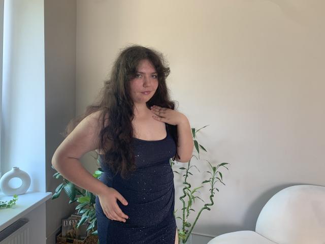 NeonilaLisova stands with long, curly brown hair, wearing a sparkly black dress, hand on hip, other hand near chest, looking at camera, in a bright room.