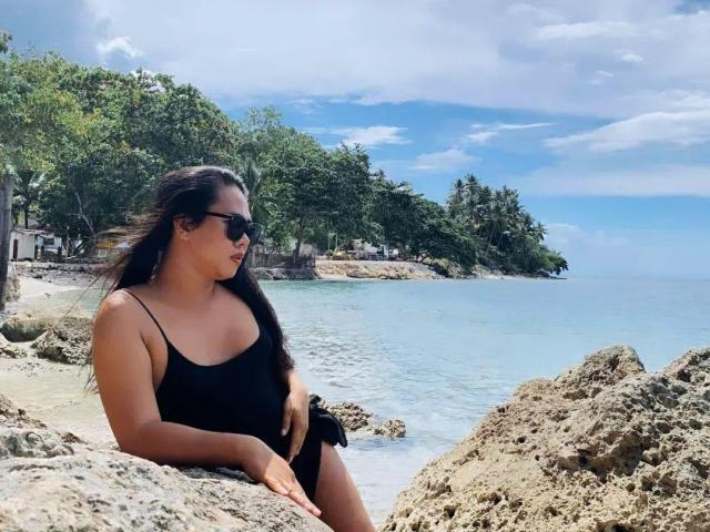 ItsmeJules with long black hair, wearing black sunglasses and a black one-piece swimsuit, sitting on rocky beach, looking to the side, with blue ocean and green trees in background.