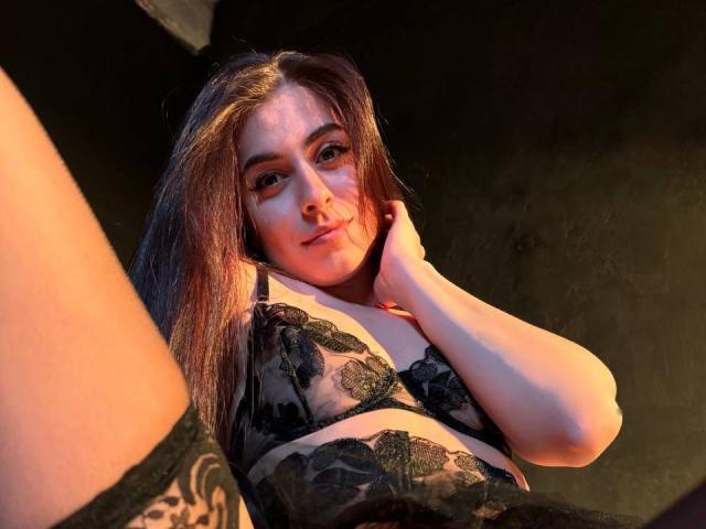 GoldBabeSi with long brown hair, wearing black lace bra, and black lace stockings, looking down with a slight smile, hand on neck, soft orange light, legs in frame.