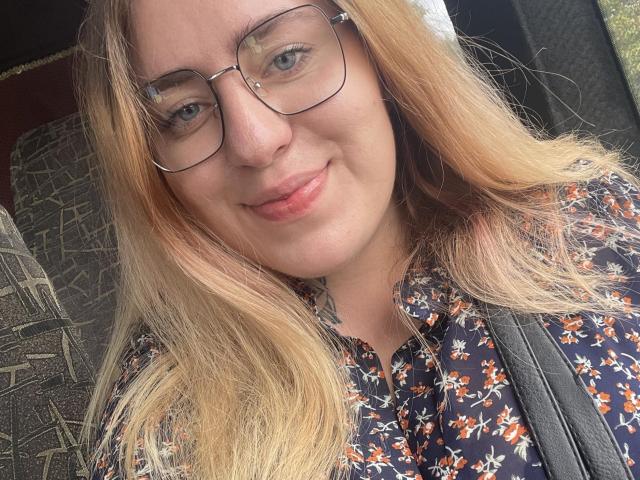 FortuneCarmen has blonde hair, glasses, and a floral shirt. She's smiling with a slight blush, showing her cute nose and pink lips. She's carrying a black bag.