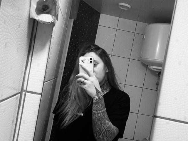 FortuneCarmen with long hair, tattoos on her right arm, taking a mirror selfie in a tiled bathroom, holding an iPhone with a floral case, wearing a black top.