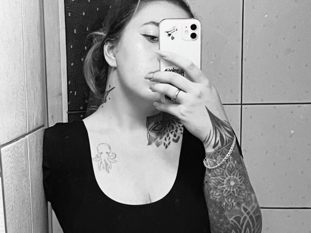 FortuneCarmen with tattoos, taking a mirror selfie with her phone, covering her mouth. She's wearing a black top, showing her chest and arm tattoos. Greyscale photo.
