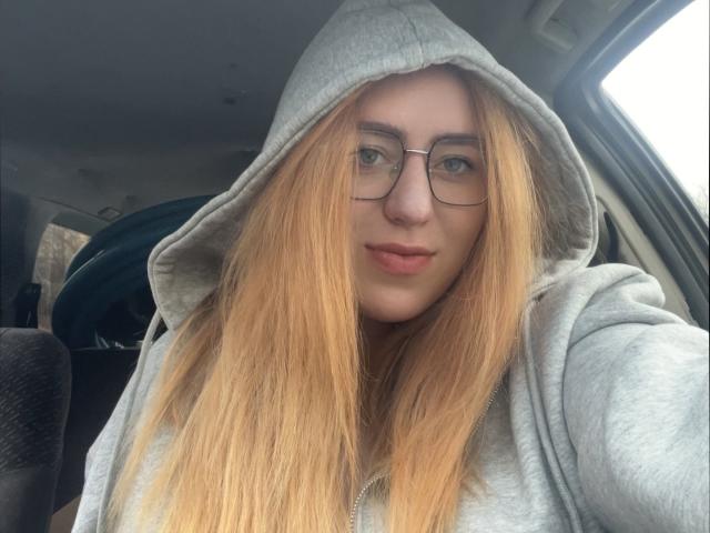 FortuneCarmen with long blonde hair, glasses, and a grey hoodie, sitting in a car, looking at the camera with a slight smile, showing her cute face and nose ring.