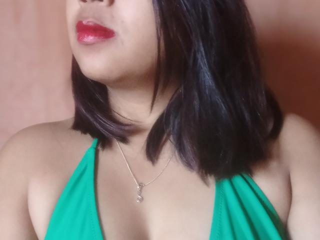 SarahFontaineX with dark brown bob, glossy red lips, green halter top, showing cleavage, wearing a silver necklace with a small pendant, against a pinkish background.