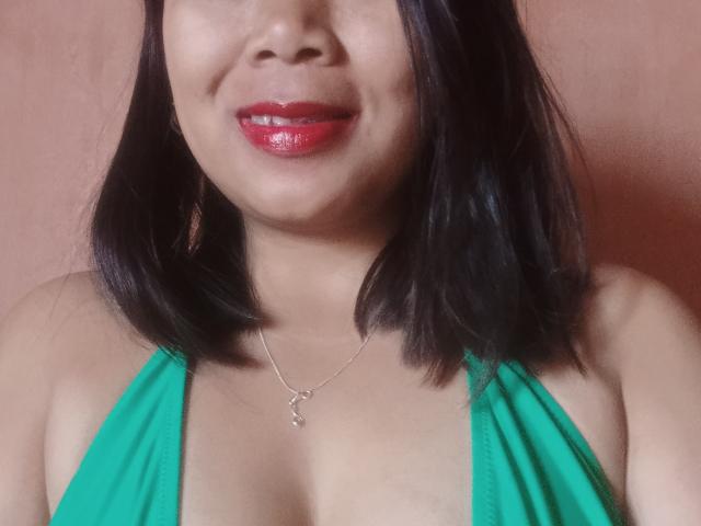 SarahFontaineX with medium brown skin, red glossy lipstick, black shoulder-length hair, green halter top, and silver necklace, showing her small tits and smiling slightly.