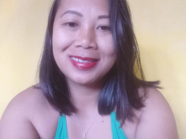 SarahFontaineX has shiny black hair, red glossy lipstick, and a teal top, showing her small tits and a cute smile with a mole above her lip.