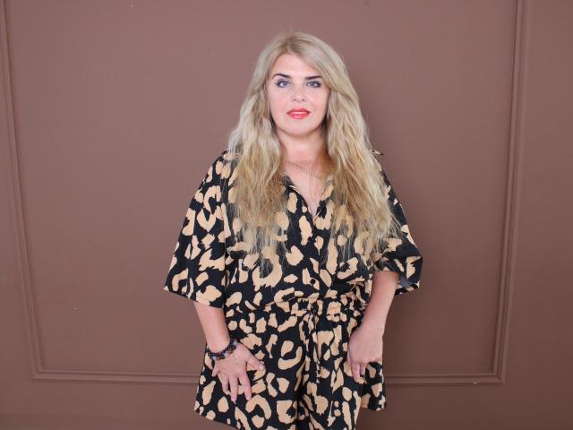 Julya Bailly, con cabello rubio largo, vestida con un vestido de estampado de leopardo en colores negro y beige, de pie frente a una pared marrón, con lápiz labial rojo y una pulsera en su muñeca derecha..