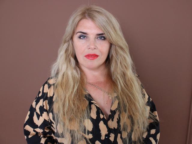 JulyaBailly has long blonde hair, red lipstick, and a black-and-beige patterned shirt. She's wearing a gold necklace against a brown background. Her tits are medium-sized.