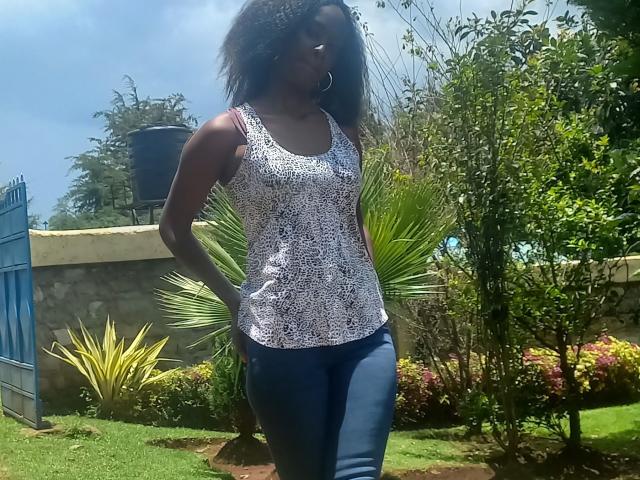 EllieNelle has dark skin, curly hair, wears a white lace tank top, blue jeans, and hoop earrings. She's standing in a sunny garden with greenery.