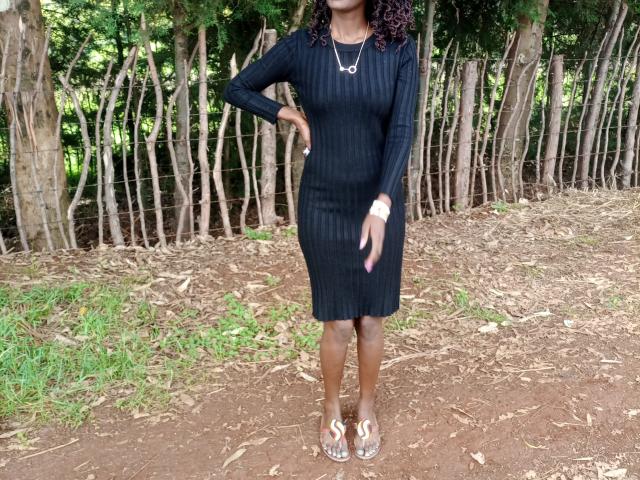 EllieNelle stands in a dirt path wearing a black ribbed dress, brown sandals, and a necklace. She has dark curly hair and a tan skin tone. Her ass is small.