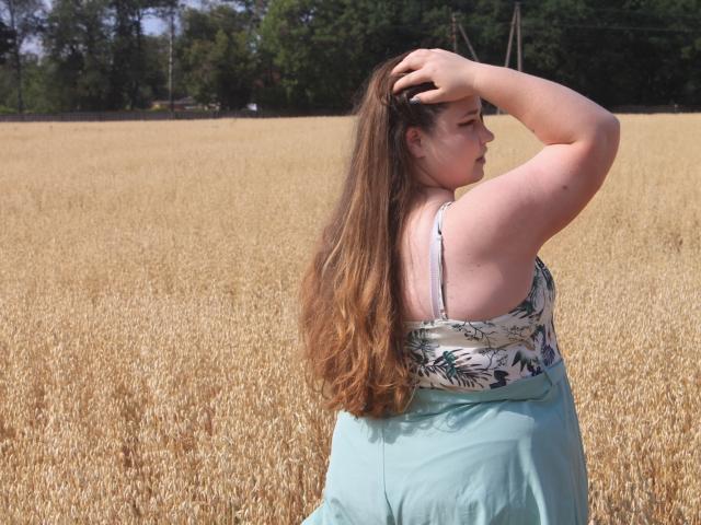 DaniellaGraceZZ with long brown hair, pale skin, wearing a floral top and green skirt, standing in a golden wheat field, raising her right arm to touch her head.
