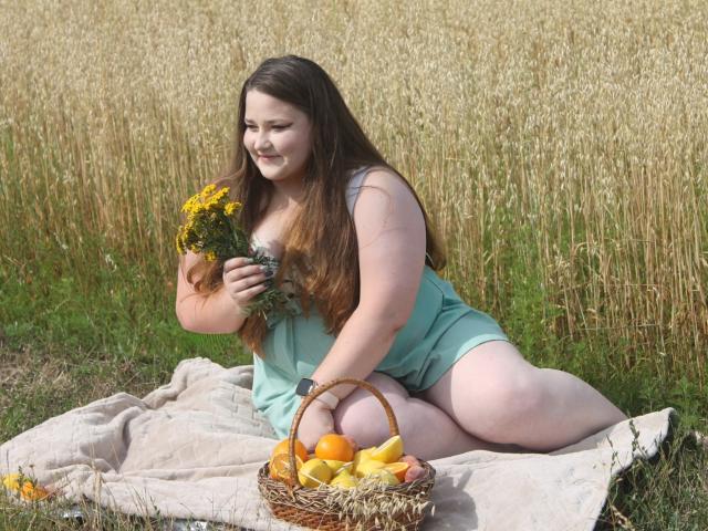 DaniellaGraceZZ, chubby with long brown hair, wearing a teal dress, sitting on a blanket, holding yellow flowers, basket with oranges and apples in front, smiling in a field.