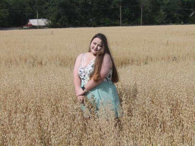 DaniellaGraceZZ stands in a wheat field, smiling, with long brown hair, wearing a white floral top and teal skirt, showing her tits slightly, in a sunny, rural setting.