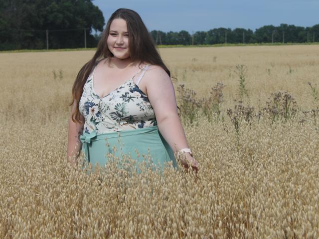 DaniellaGraceZZ stands in a golden wheat field, smiling with long brown hair, wearing a floral top and green skirt, showing her boobs and ass slightly hidden by the wheat.
