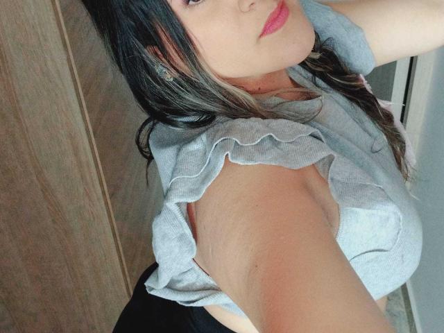 MiaJhonss tem pele clara, batom rosa e cabelo castanho. Ela está vestindo uma blusa cinza com babados, que mostra seus seios e axilas. Ela está tirando uma selfie, olhando levemente para cima..