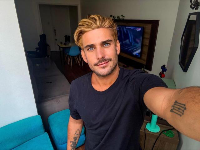SurferKing, blonde dude with blue eyes, black t-shirt, tattoos on arms, mustache, taking selfie, blue couch in background, green dildo on wooden table, smiling, slightly muscular.