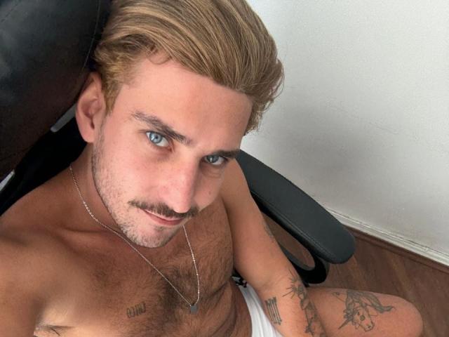 SurferKing with blonde hair, blue eyes, and a mustache, wearing white underwear, sitting in a chair, chest tattoo, arm tattoos, wooden floor, and a silver necklace.
