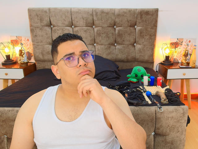 DmitriIvanov, chubby dude with short black hair, purple glasses, white tank top, thinking with finger on chin, bed with tufted brown headboard, sex toys, green plushie, and candles.