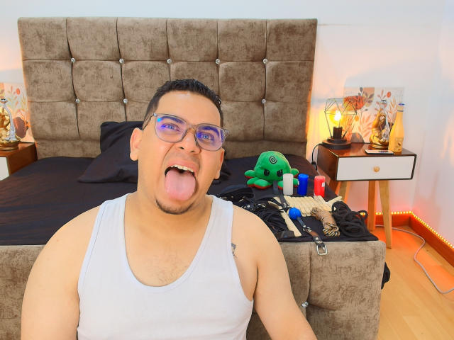 DmitriIvanov, chubby dude with glasses, white tank top, tongue out, short black hair, bed with brown tufted headboard, sex toys on nightstand, green stuffed toy, bedside lamp.