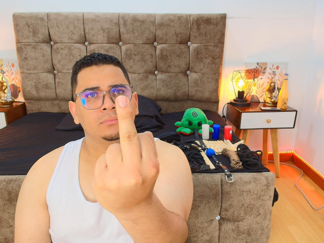 DmitriIvanov, a chubby guy with short dark hair and glasses, points a finger at the camera with a serious expression, wearing a white tank top, in a bedroom.