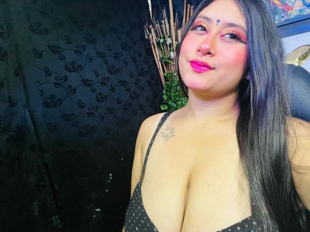 KalpanaAisha with pink eyeshadow, black hair, and pink lipstick, showing her big tits in a black polka dot top, smiling at the camera. Tattoo on her right chest.