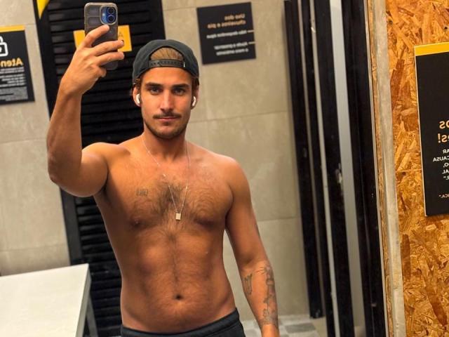 Shirtless SurferKing with a lean, hairy chest, wearing a black cap, taking a mirror selfie, showing a tattoo on his right arm, wearing dark pants, and a necklace.