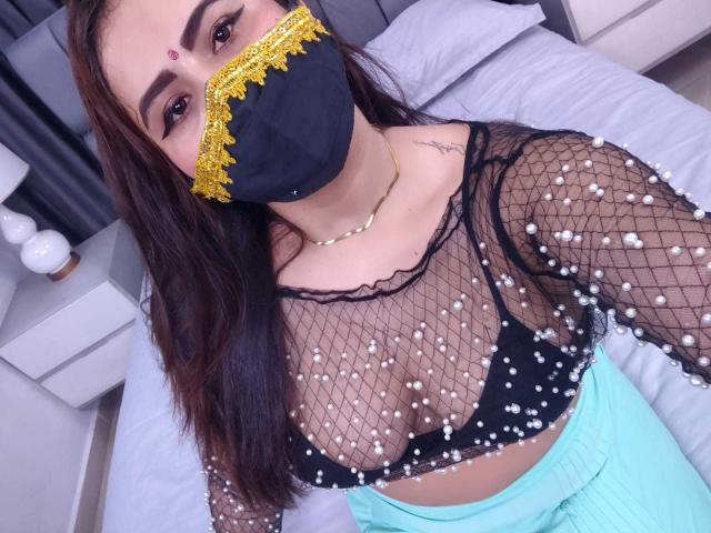 DaliaDubey with dark brown hair, wearing a black mesh top with white pearls, black mask with yellow lace, and teal pants, showing her tits and gold necklace.