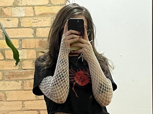 AmySpider, con cabello largo y castaño, vestida con una camiseta negra con un estampado de araña roja, mangas de red en los brazos, y sosteniendo un teléfono, cubriendo su rostro..