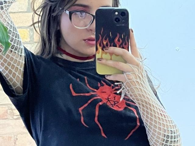 AmySpider with glasses, wearing a black Spider t-shirt, fishnet arms, red choker, taking a mirror selfie with a flame-patterned phone, wet brown hair, light skin, slight pout.