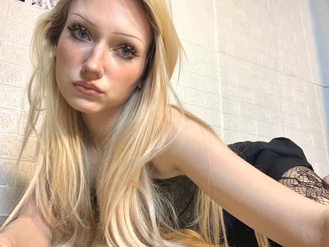 AngelTemptress69 has long blonde hair, blue eyes, and a serious expression, wearing a black lace top, showing her bare shoulders and upper chest, in a selfie.