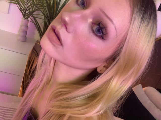 AngelTemptress69 has shiny pink-tinted skin, blue eyes, and blonde hair. Her lips are slightly parted, showing a subtle, sexy expression. She's looking up with a slight tilt.