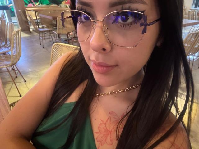 AlisonTatcher with glasses, green top, and chain necklace, showing her tits with a floral tattoo, smiling, dark hair, and blue eyes, sitting in a café.