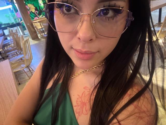 Alison Tatcher, con gli occhiali, indossa una maglietta verde, ha i capelli neri, una catena d'oro, un tatuaggio floreale rosso sulla spalla, sta sorridendo, ha un seno di media grandezza, pelle chiara, naso visibile, labbra rosa pallido, si trova in un accogliente caffè..