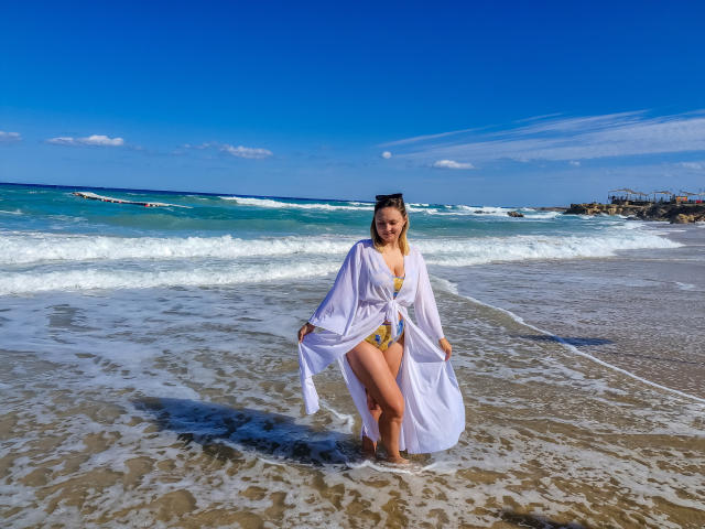 SallyVerda in a white robe, yellow bikini, standing in shallow ocean water, waves crashing, blue sky, white clouds, sandy beach, blonde hair, smiling, holding robe, legs together.
