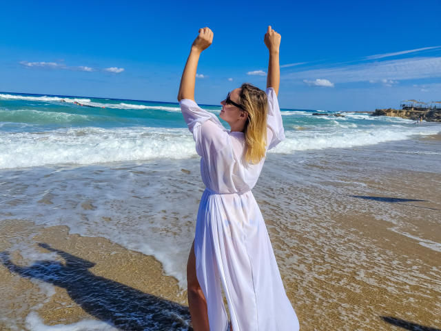 SallyVerda stands on a beach, arms raised, wearing a white, sheer dress, showing a thigh, with blonde hair, sunglasses, and a blue sky.