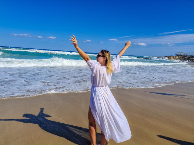SallyVerda with arms raised, wearing a white dress, standing on a beach, blue sky, ocean, and waves, shadow on sand, blonde hair, sunglasses, sunny day, sexy.